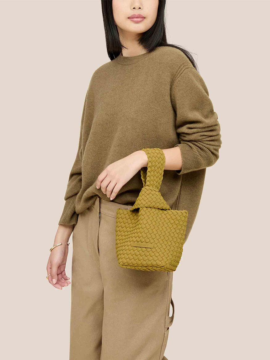 A person wearing a brown sweater and beige pants holds a Naghedi Kyoto Clutch in Solid Chartreuse, adding a touch of elegance to evening outfits with its handwoven neoprene design.