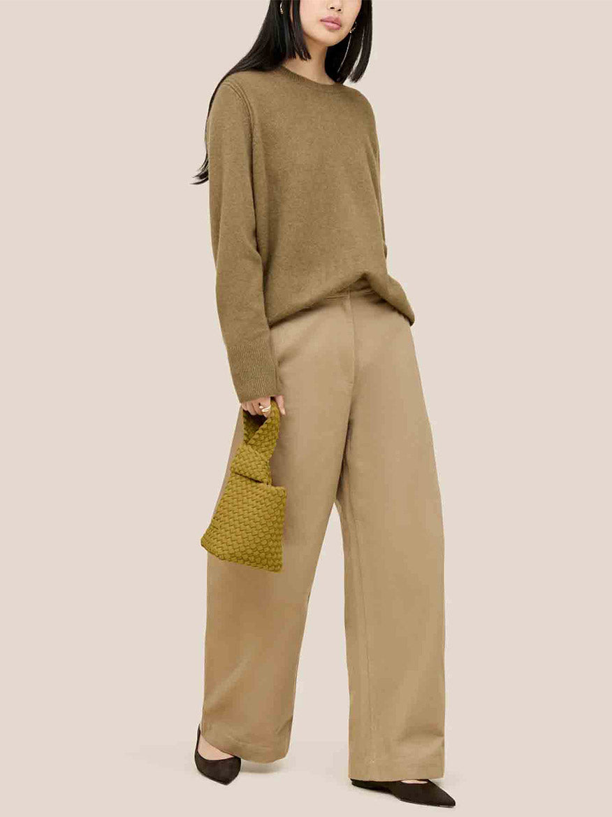 A woman in a brown sweater and wide-legged pants holds a chartreuse handbag that captures the essence of a Naghedi Kyoto Clutch, creating an elegant silhouette against a plain beige background, perfect for chic evening ensembles.