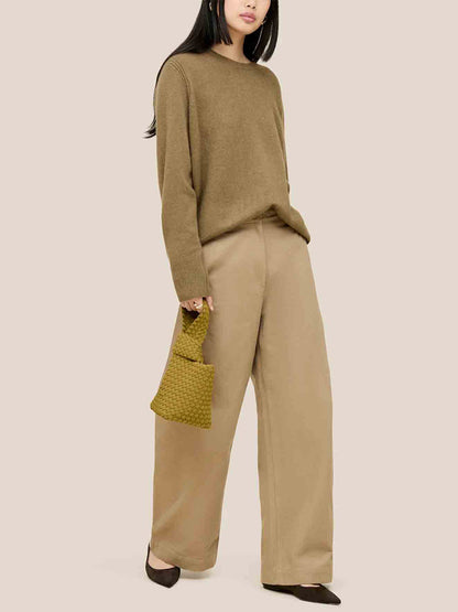 A woman in a brown sweater and wide-legged pants holds a chartreuse handbag that captures the essence of a Naghedi Kyoto Clutch, creating an elegant silhouette against a plain beige background, perfect for chic evening ensembles.