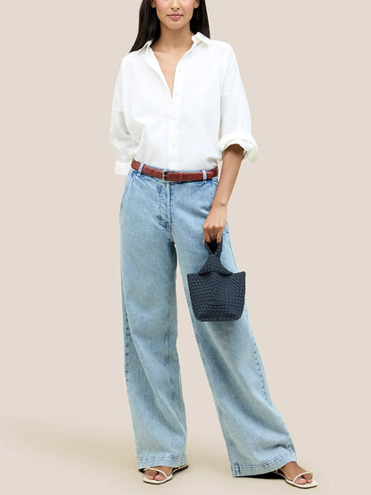 A person wearing a white shirt and blue wide-leg jeans holds the Naghedi Kyoto Clutch in Solid Murano, a small woven bag perfect for evening outfits.