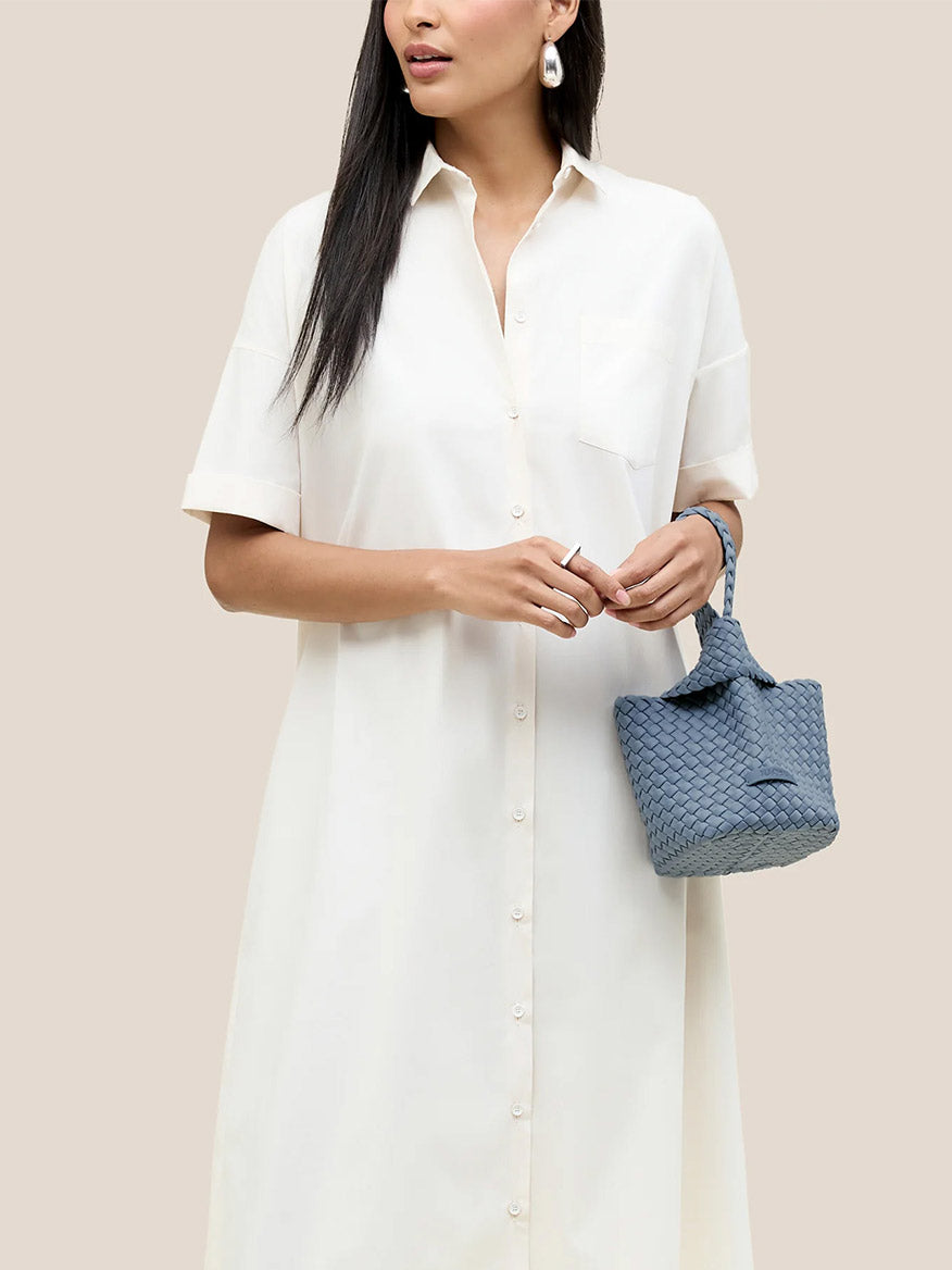 A person in a white shirt dress holds the Naghedi Kyoto Clutch in Solid Nova, standing against a plain background, perfect for elegant evening outfits.