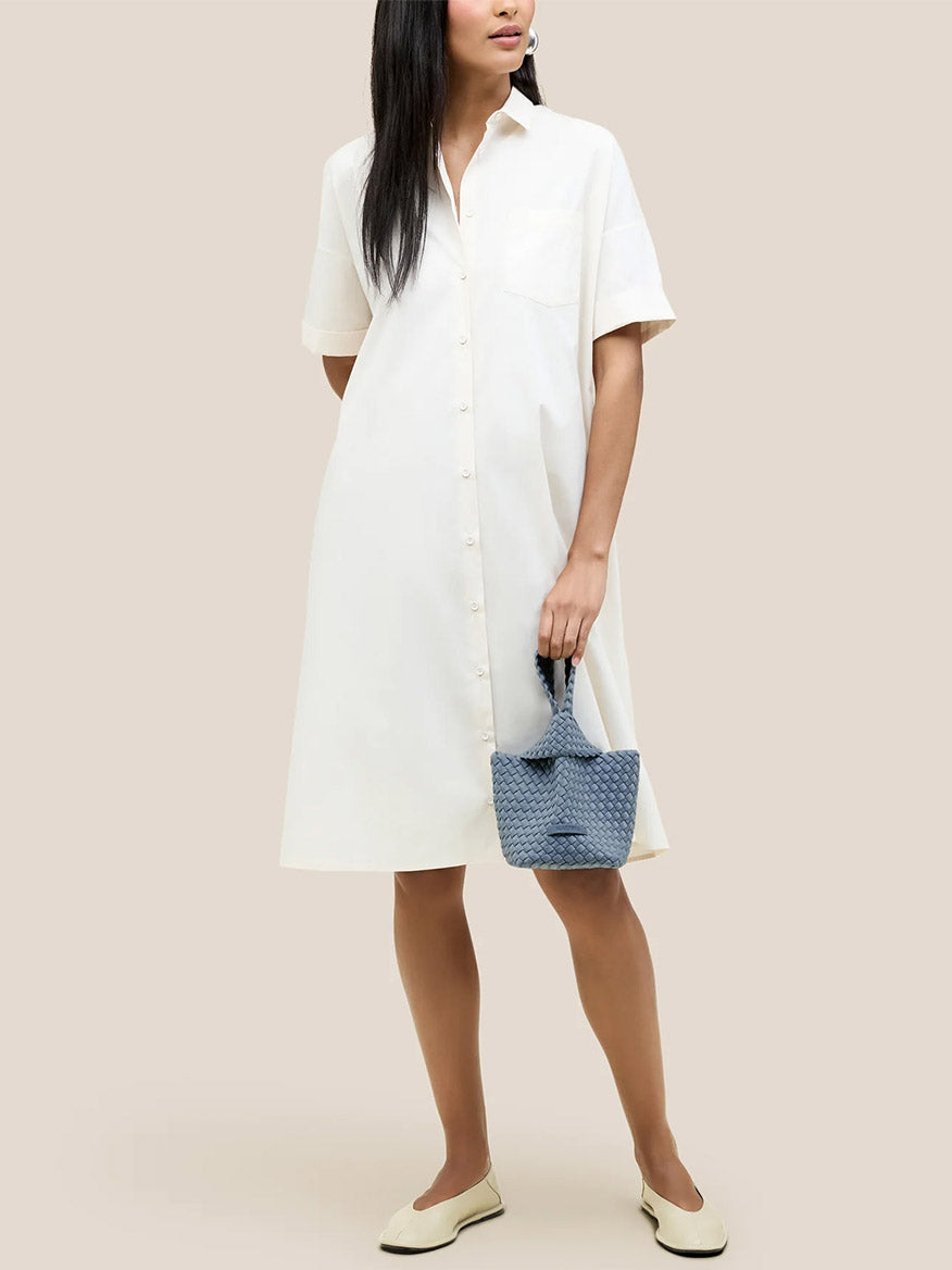 A person wearing a white button-up dress, holding a small blue Naghedi Kyoto Clutch in Solid Nova, and wearing white shoes against a beige background.