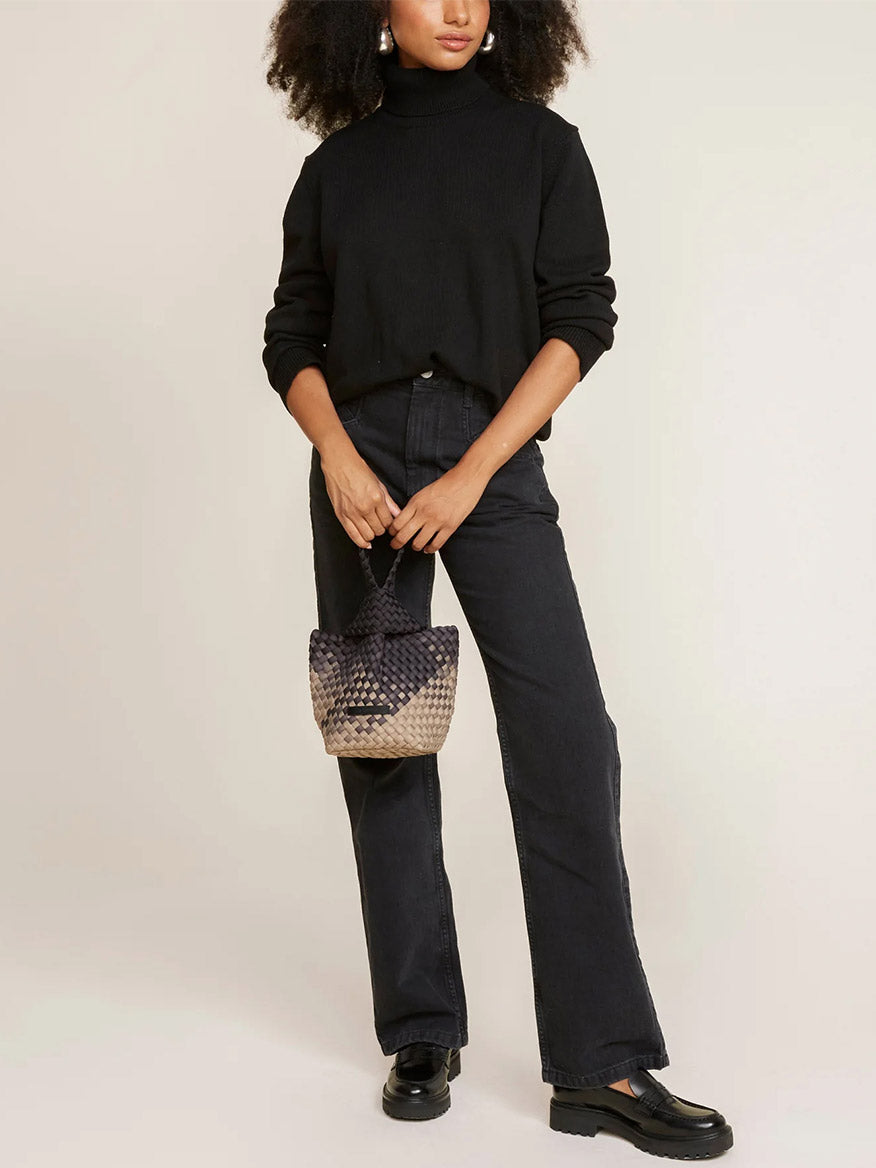 Person wearing a black turtleneck sweater, dark jeans, and loafers, holding a Naghedi Kyoto Clutch in Graphic Ombre Mahal, standing against a beige background.
