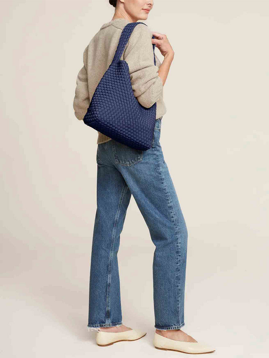 A person in jeans and a beige sweater stands sideways with the spacious and comfortable Naghedi Nomad Hobo in Solid Ink Blue, crafted from handwoven neoprene, slung over their shoulder.