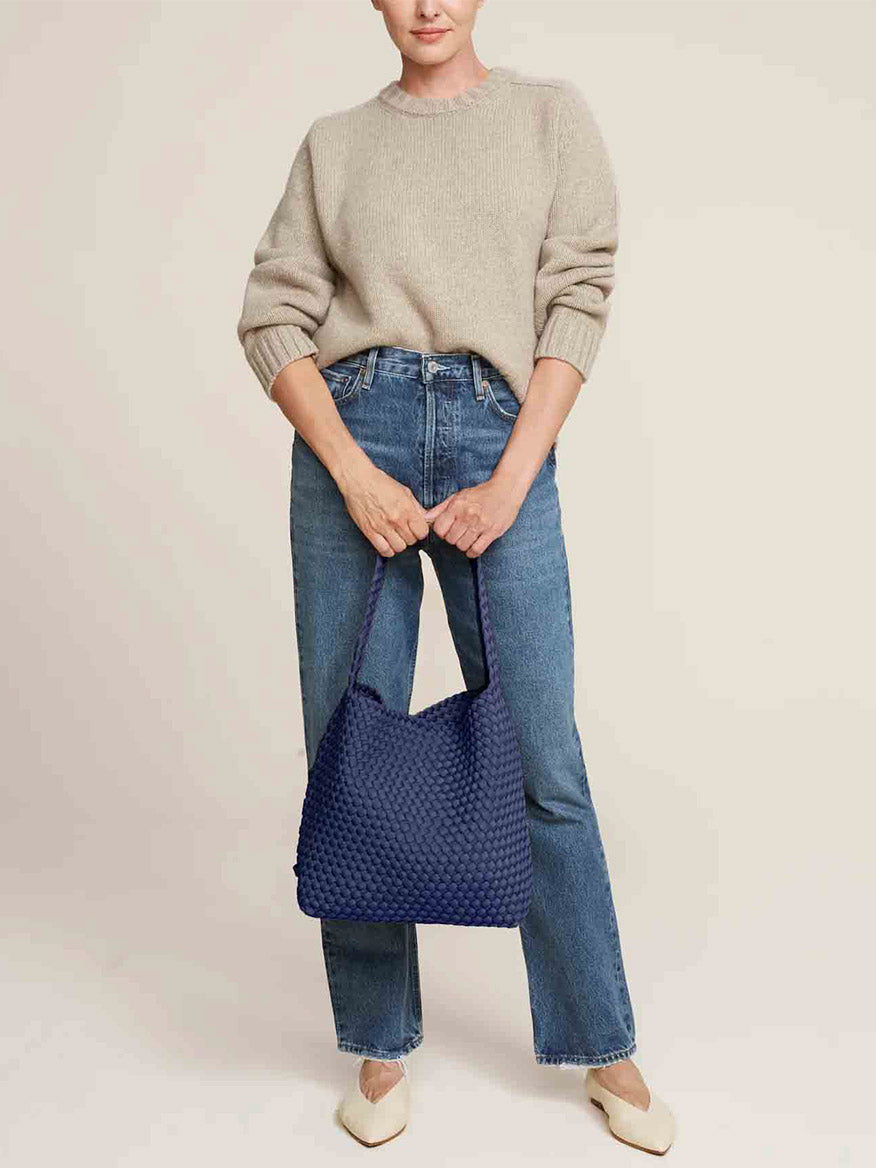 A person in a beige sweater and blue jeans holds a spacious Naghedi Nomad Hobo in Solid Ink Blue, its handwoven neoprene design making a stylish statement.