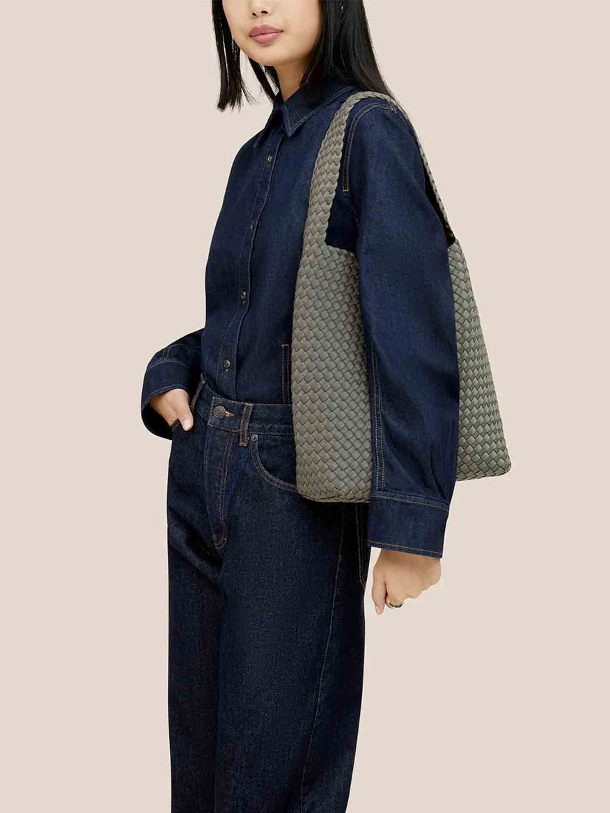 Dressed in dark denim, a person carries the roomy and comfortable Naghedi Nomad Hobo in Solid Laurel, a textured gray shoulder bag made from handwoven neoprene, set against a neutral background.