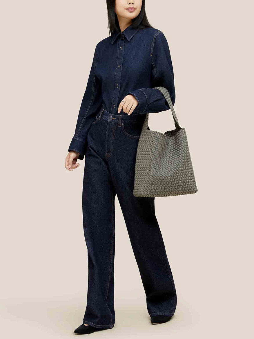 A person wearing a dark denim shirt and jeans holds a textured gray handbag, expertly crafted in handwoven neoprene, embodying the essence of the Naghedi Nomad Hobo in Solid Laurel—spacious and comfortable for any journey.