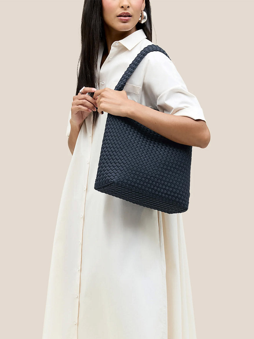 A person in a white dress carries the Naghedi Nomad Hobo in Solid Murano, featuring its handwoven neoprene design, on their shoulder against a plain background.