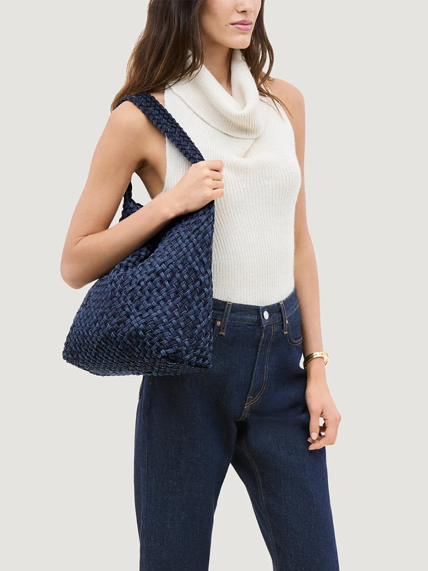 A woman in a sleeveless cream turtleneck and high-waisted blue jeans carries the Naghedi Nomad Medium Hobo Double Weave Denim bag in Indigo.