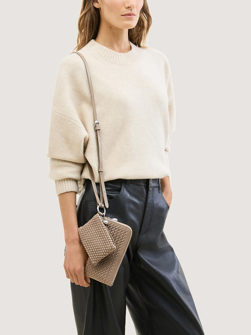 A woman in a beige sweater and black trousers wearing a Naghedi Orly Phone Case in Cashmere as a beige woven crossbody phone bag with a matching small accessory.