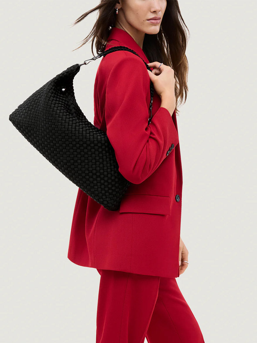 A woman in a red suit holds the Naghedi Porto Medium Half-Moon Bag in Onyx against a plain light background.