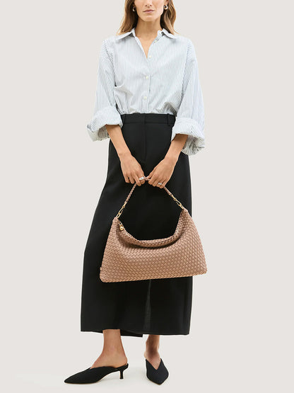 A person holds a crescent-shaped, woven tan Naghedi Porto Medium Half-Moon Bag in Seychelles made of neoprene with a gold-tone chain handle.