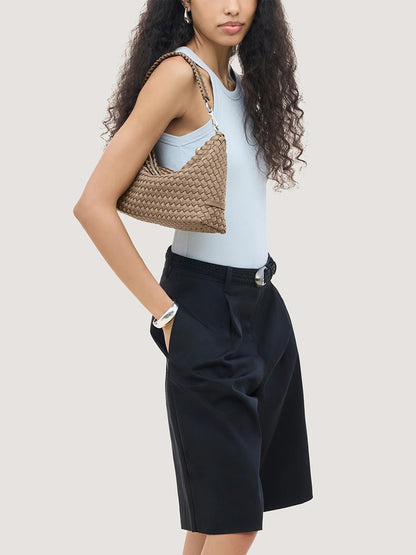 A person wearing a light blue tank top and black trousers carries a tan, handwoven neoprene Naghedi Porto Medium Half-Moon Bag in Cashmere.
