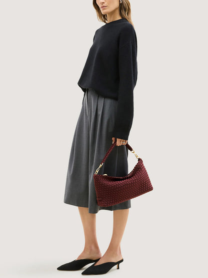 A person carrying a Naghedi Porto Small Half-Moon Bag in bordeaux, featuring a handwoven crescent shape, braided strap, and gold-tone hardware.