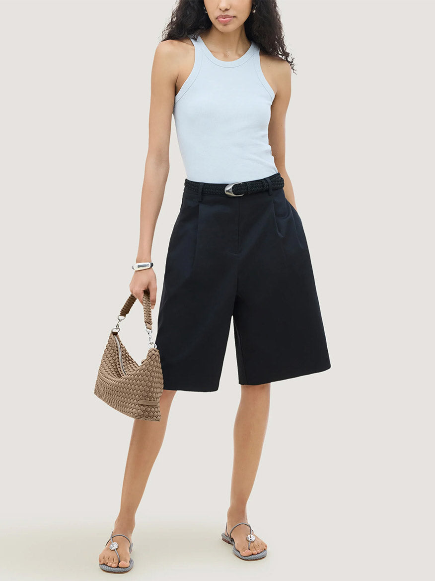 A person wearing a light blue tank top and dark shorts holds a handwoven neoprene Naghedi Porto Small Half-Moon Bag in Cashmere.