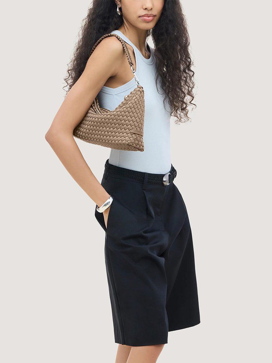 A person in a light blue tank top and black trousers carries a brown, handwoven Naghedi Porto Small Half-Moon Bag.