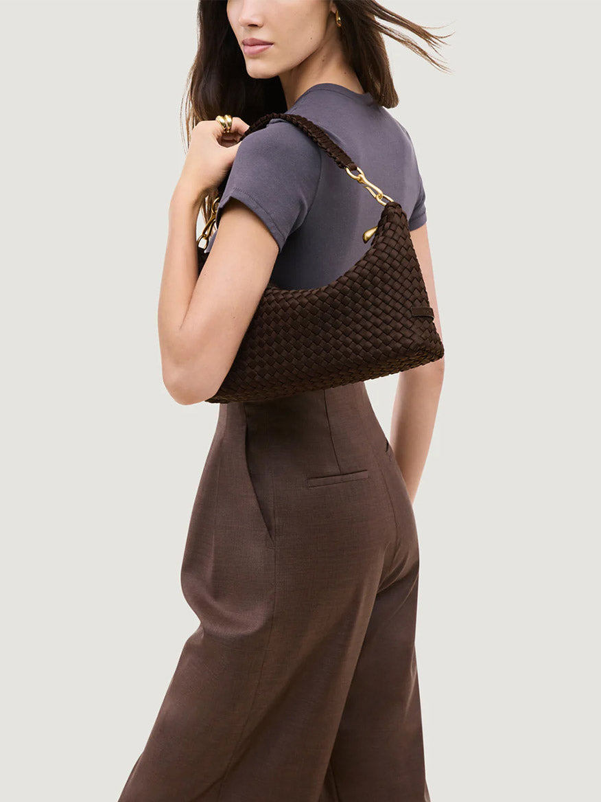Woman in a short-sleeve grey top and brown pants carries the Naghedi Porto Small Half-Moon Bag in Chocolate, featuring gold hardware, over her shoulder against a plain background.