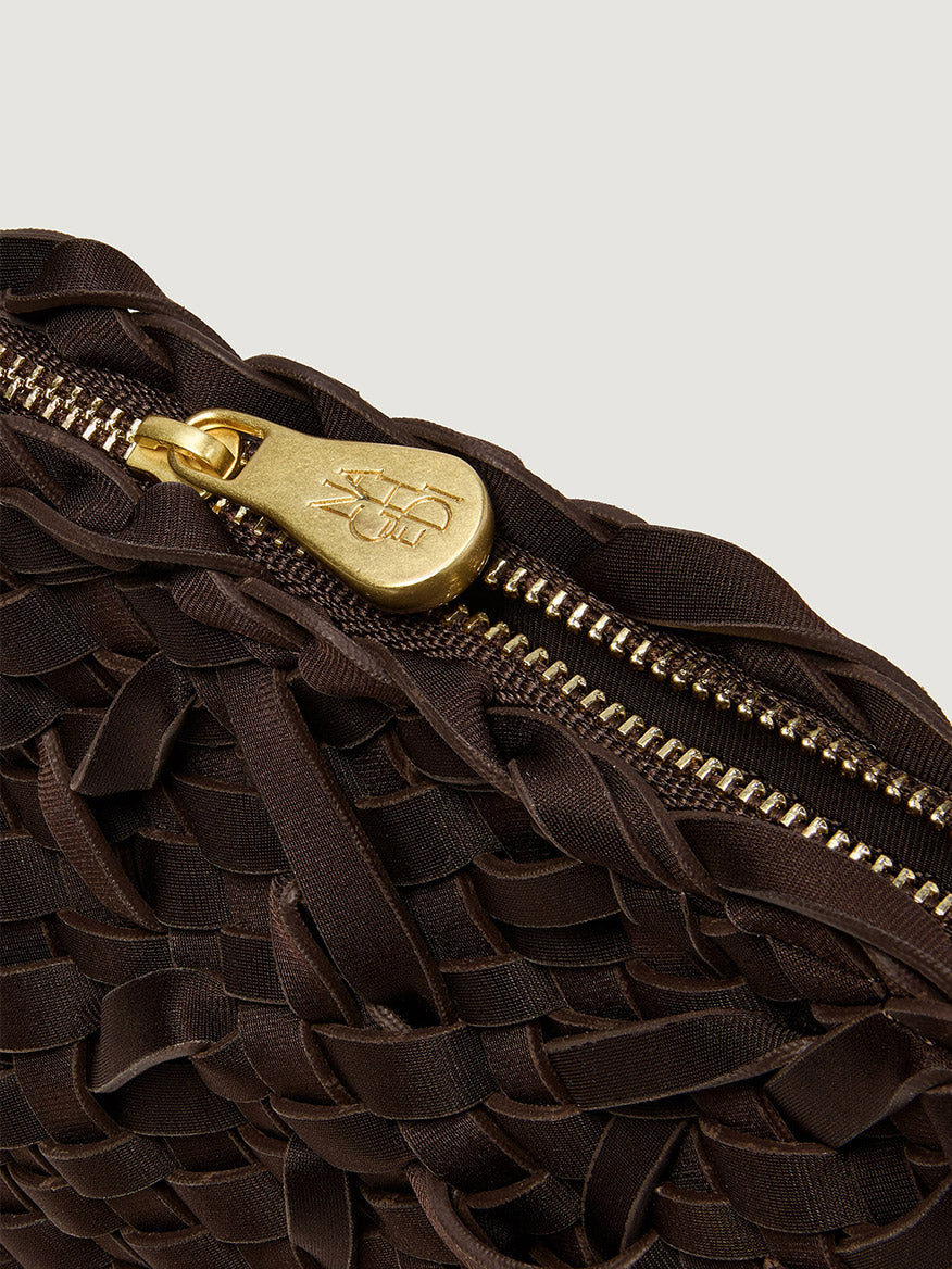 Close-up of a dark brown Naghedi Soho Crossbody Nest Weave in Chocolate, a handwoven neoprene bag featuring a gold zipper pull with embossed logo lettering.
