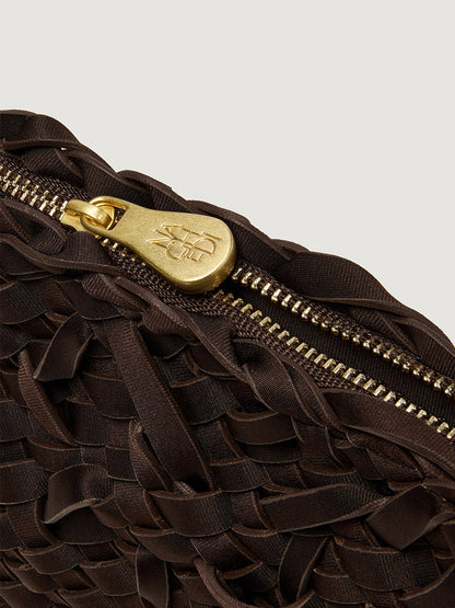 Close-up of a dark brown Naghedi Soho Crossbody Nest Weave in Chocolate, a handwoven neoprene bag featuring a gold zipper pull with embossed logo lettering.