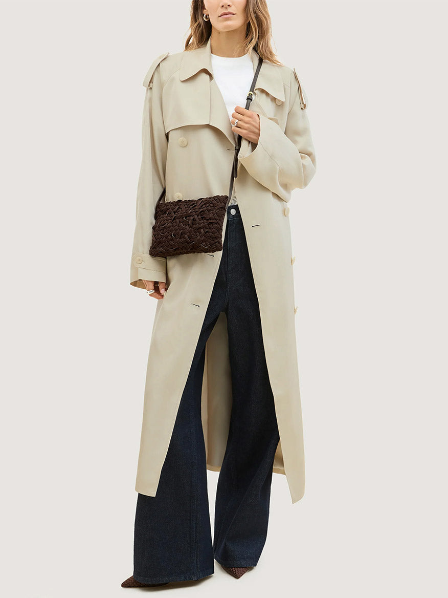 A person wearing a long beige trench coat and dark flared jeans carries a dark brown Naghedi Soho Crossbody Nest Weave in Chocolate.