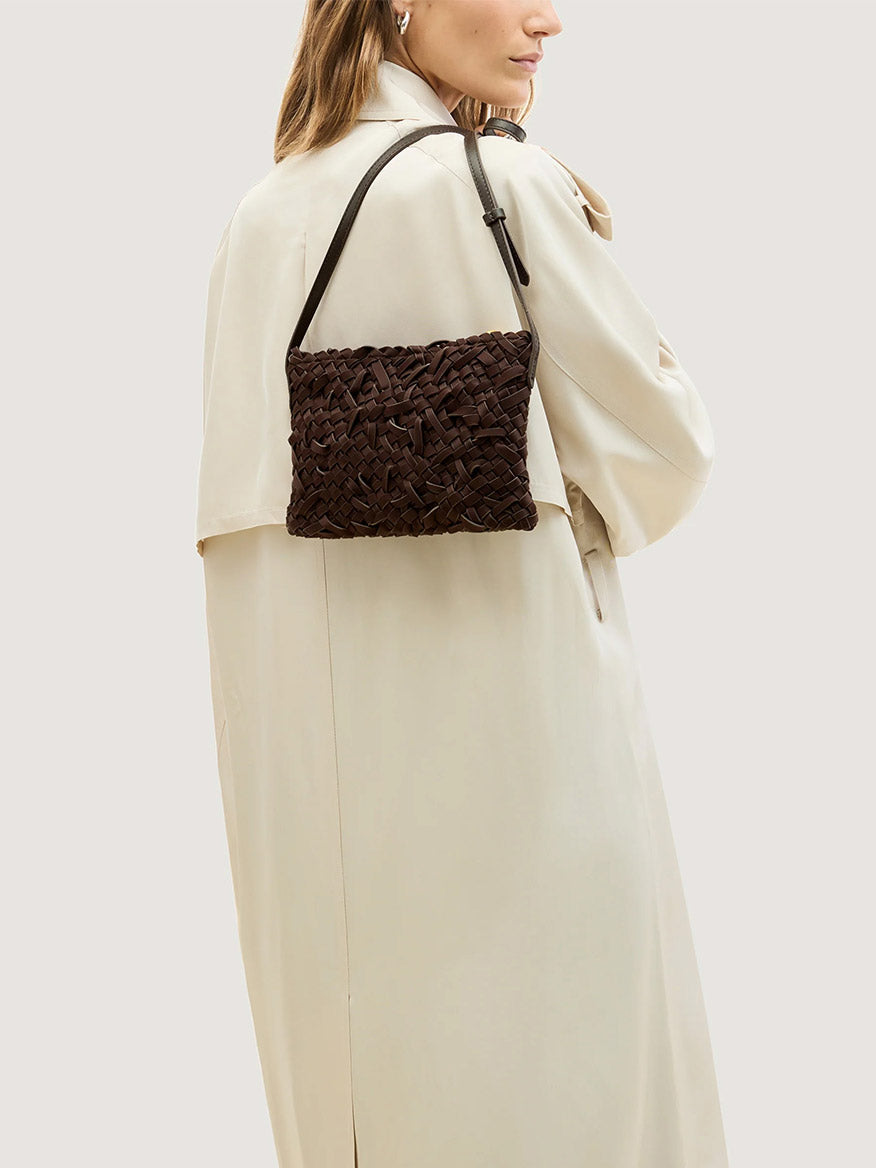 A person wearing a beige trench coat carries a dark brown Naghedi Soho Crossbody Nest Weave in Chocolate shoulder bag.