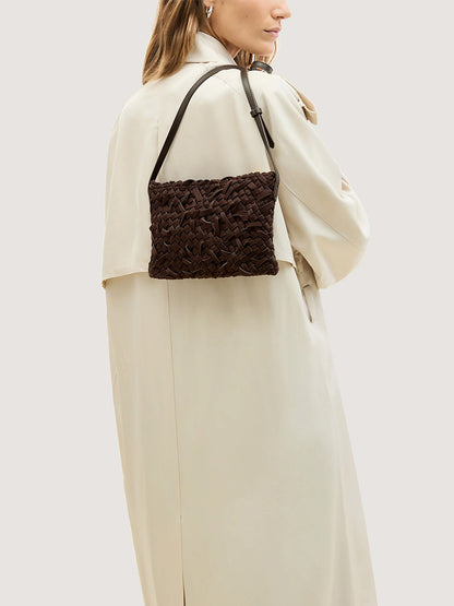 A person wearing a beige trench coat carries a dark brown Naghedi Soho Crossbody Nest Weave in Chocolate shoulder bag.