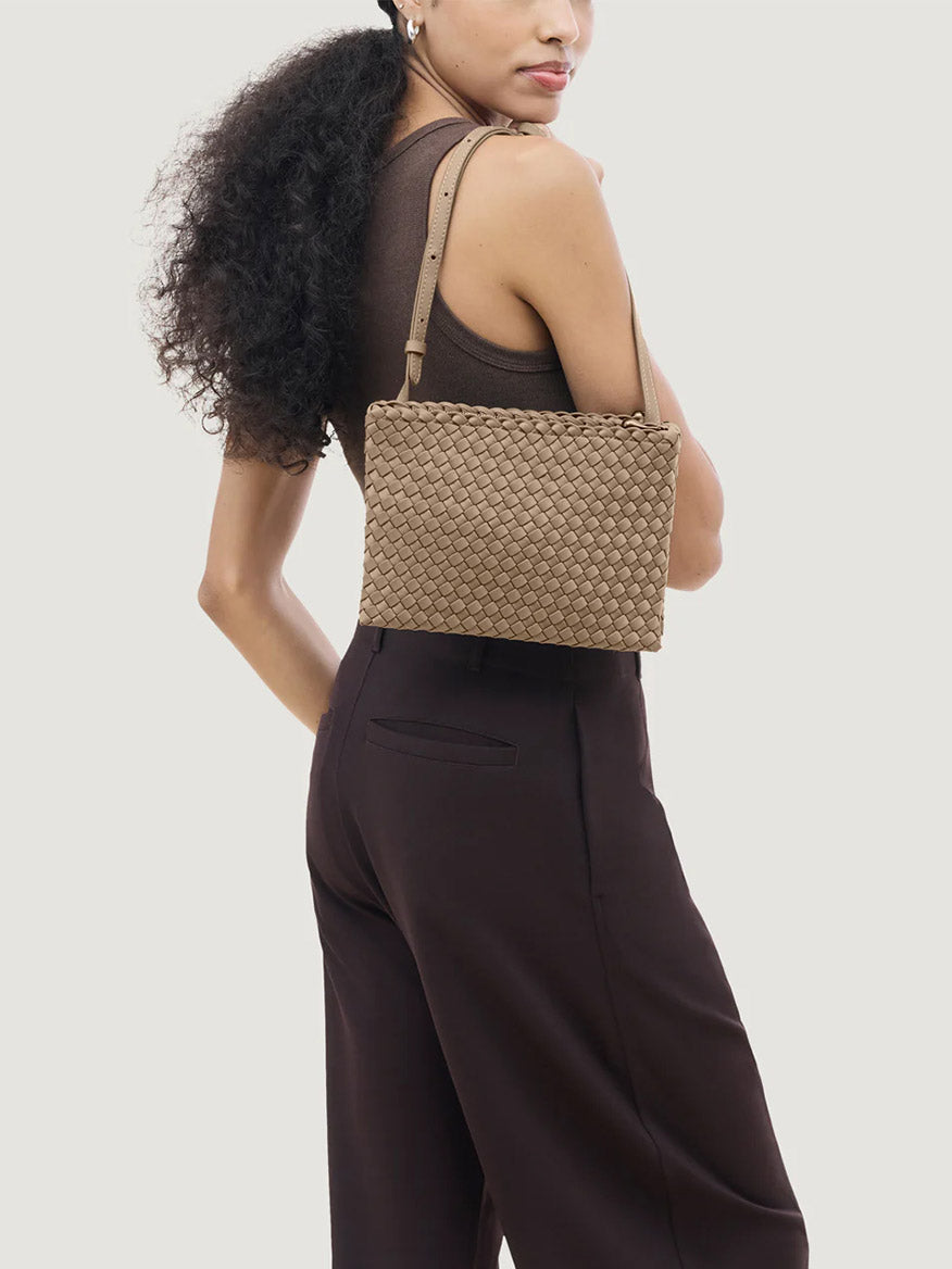 A person wears a brown sleeveless top and dark pants, carrying the Naghedi Soho Envelope Crossbody in Cashmere with an adjustable leather strap, posing against a plain light background.