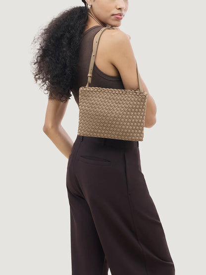 A person wears a brown sleeveless top and dark pants, carrying the Naghedi Soho Envelope Crossbody in Cashmere with an adjustable leather strap, posing against a plain light background.