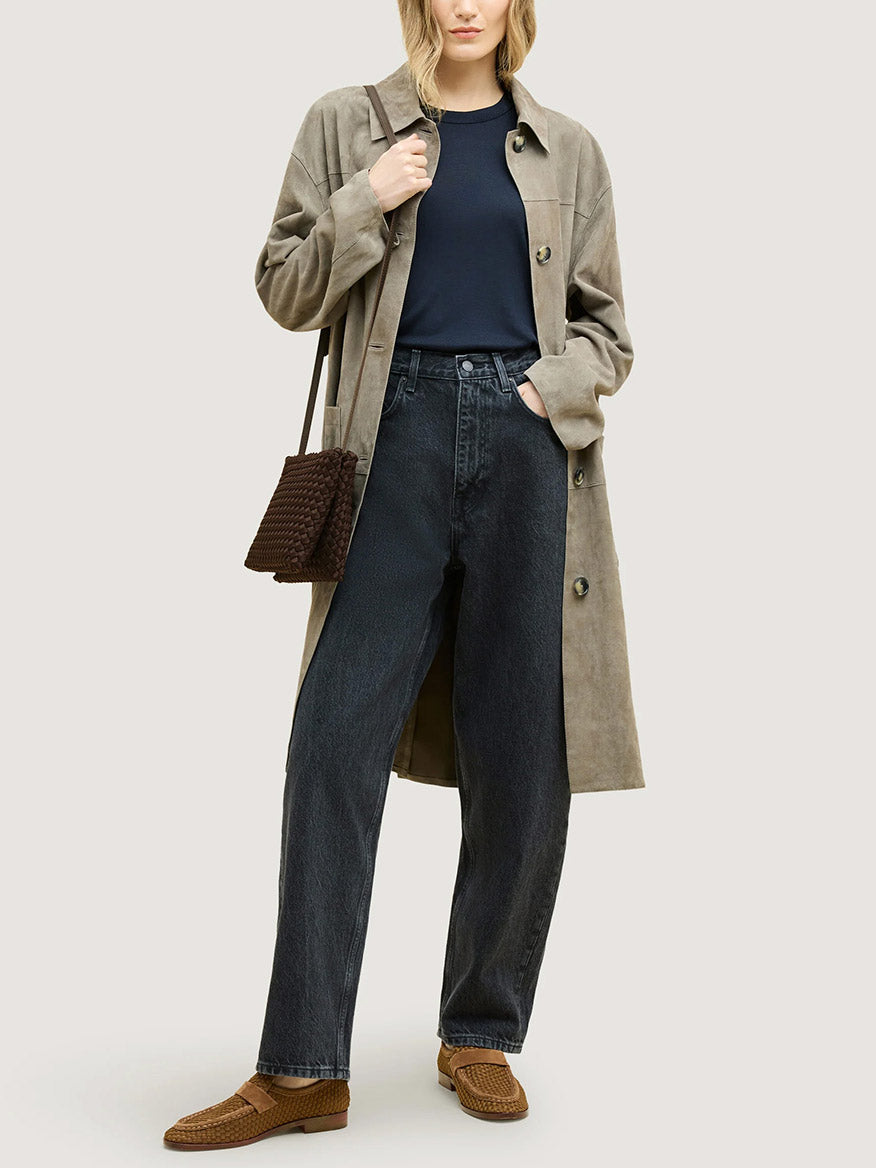A person wearing a long jacket, dark shirt, and jeans, carrying a chocolate Naghedi Soho Envelope Crossbody in a small, dark brown woven neoprene design with an adjustable strap.