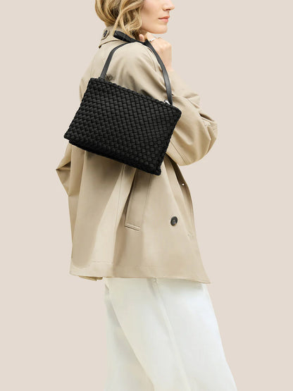 A woman in a beige trench coat and white pants carries the Naghedi Soho Envelope Crossbody in Onyx, a black woven neoprene bag with an adjustable strap, over her shoulder against a plain light background.