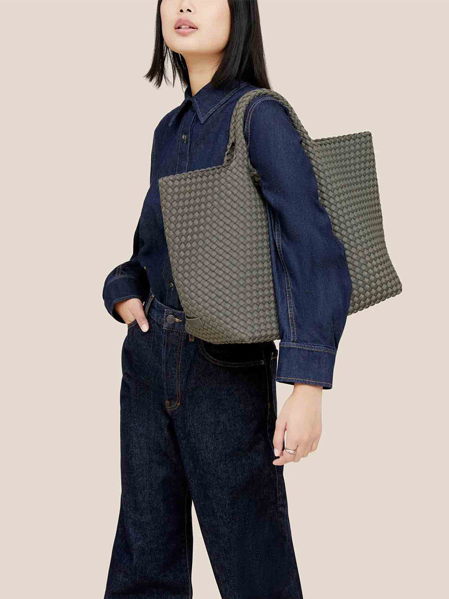 Dressed in a dark denim outfit, a person carries the Naghedi St. Barths Large Tote in Solid Laurel over their shoulder, its solid laurel texture complemented by a spacious shoulder strap.