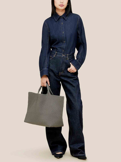 A person in a dark denim outfit carries the Naghedi St. Barths Large Tote in Solid Laurel with its handwoven neoprene design, featuring a spacious shoulder strap, against a plain background.