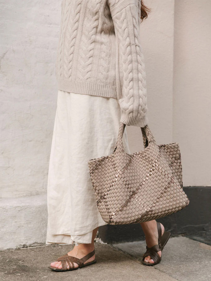 Woman in a cable knit sweater and white skirt carrying a Naghedi St. Barths Medium Tote in Graphic Weave Palermo, standing against a white wall.
