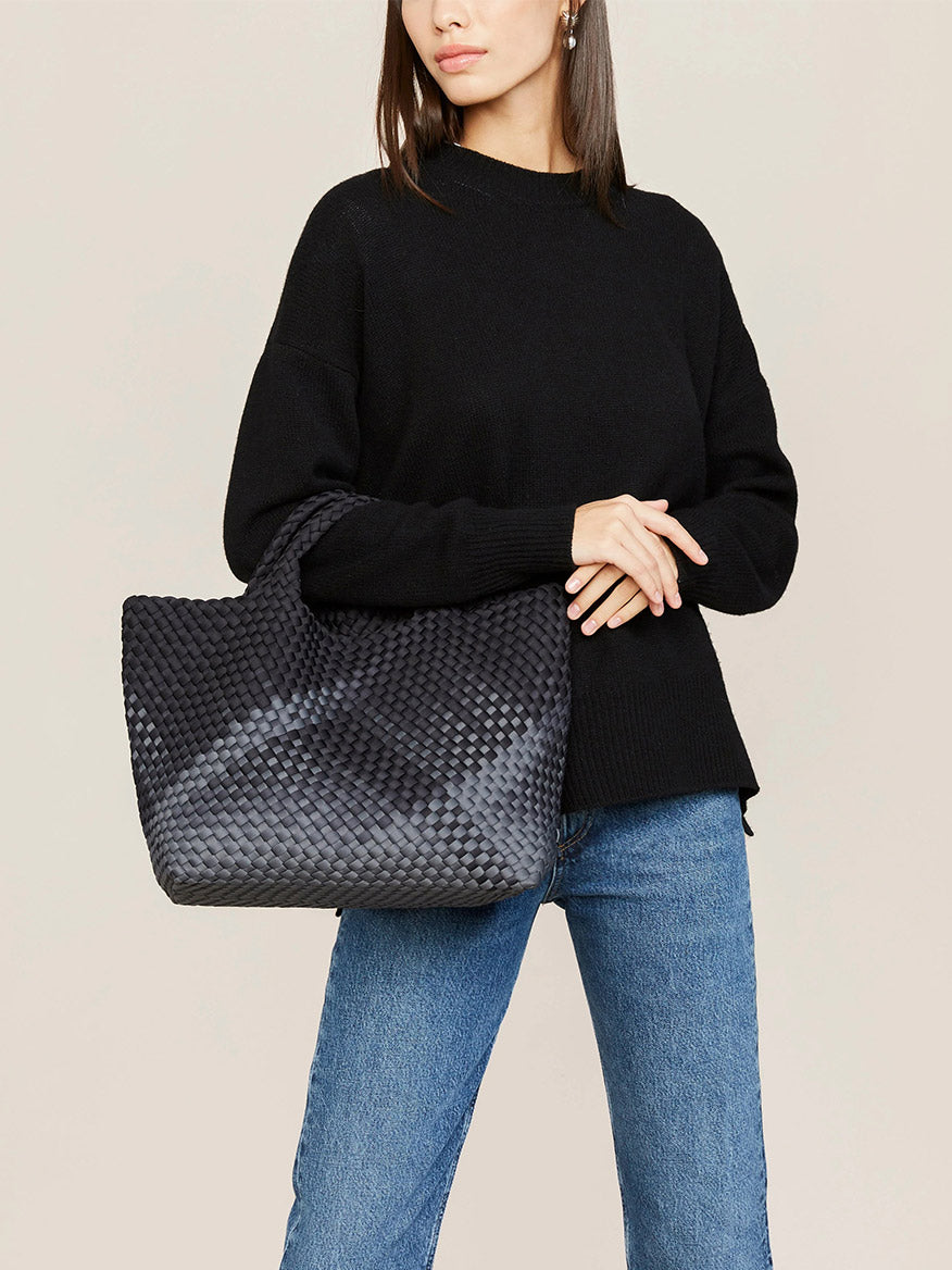 A woman wearing a black sweater and jeans holding a Naghedi St. Barths Medium Tote in Graphic Ombre Basalt.