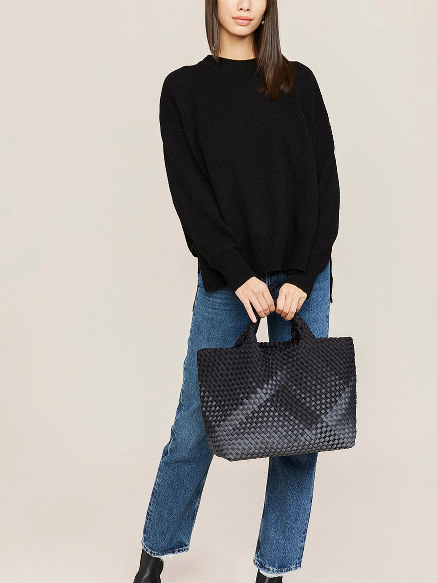 The model is wearing a black sweater and jeans with a Naghedi St. Barths Medium Tote in Graphic Ombre Basalt.