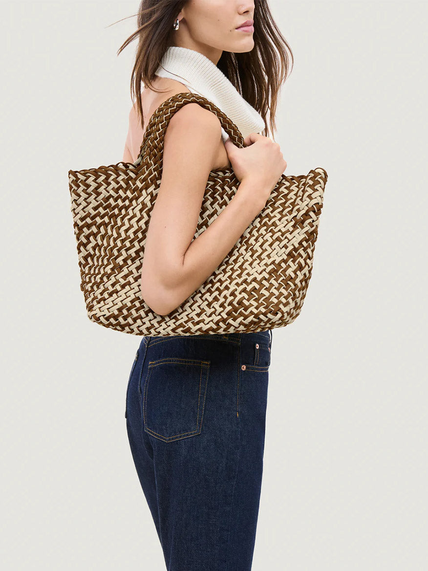 A woman in dark jeans and a sleeveless top carries the Naghedi St. Barths Medium Tote in Double Weave Carmel with gold hardware over her shoulder against a plain background.
