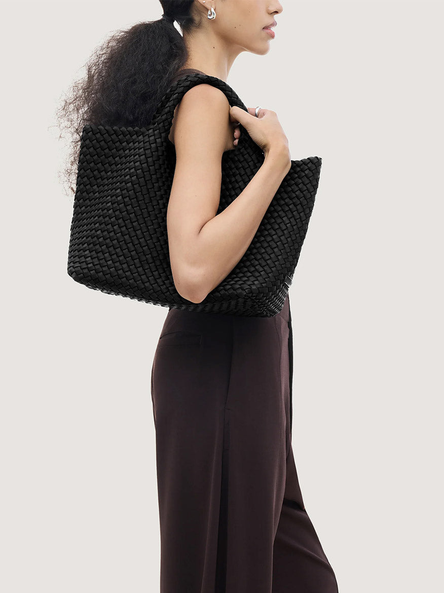A person carrying a black woven Naghedi St. Barths Medium Tote in Onyx over their shoulder.