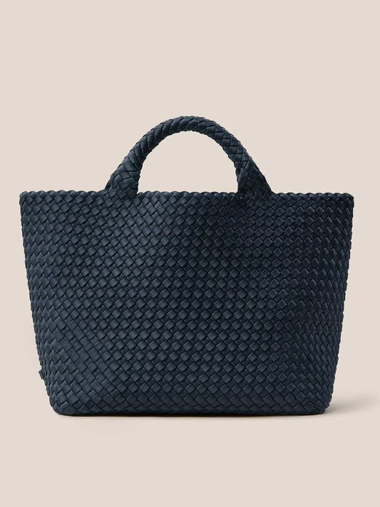 The Naghedi St. Barths Medium Tote in Solid Murano, a dark blue handwoven neoprene bag with two short handles, stands out beautifully against a beige background, making it the perfect everyday bag.