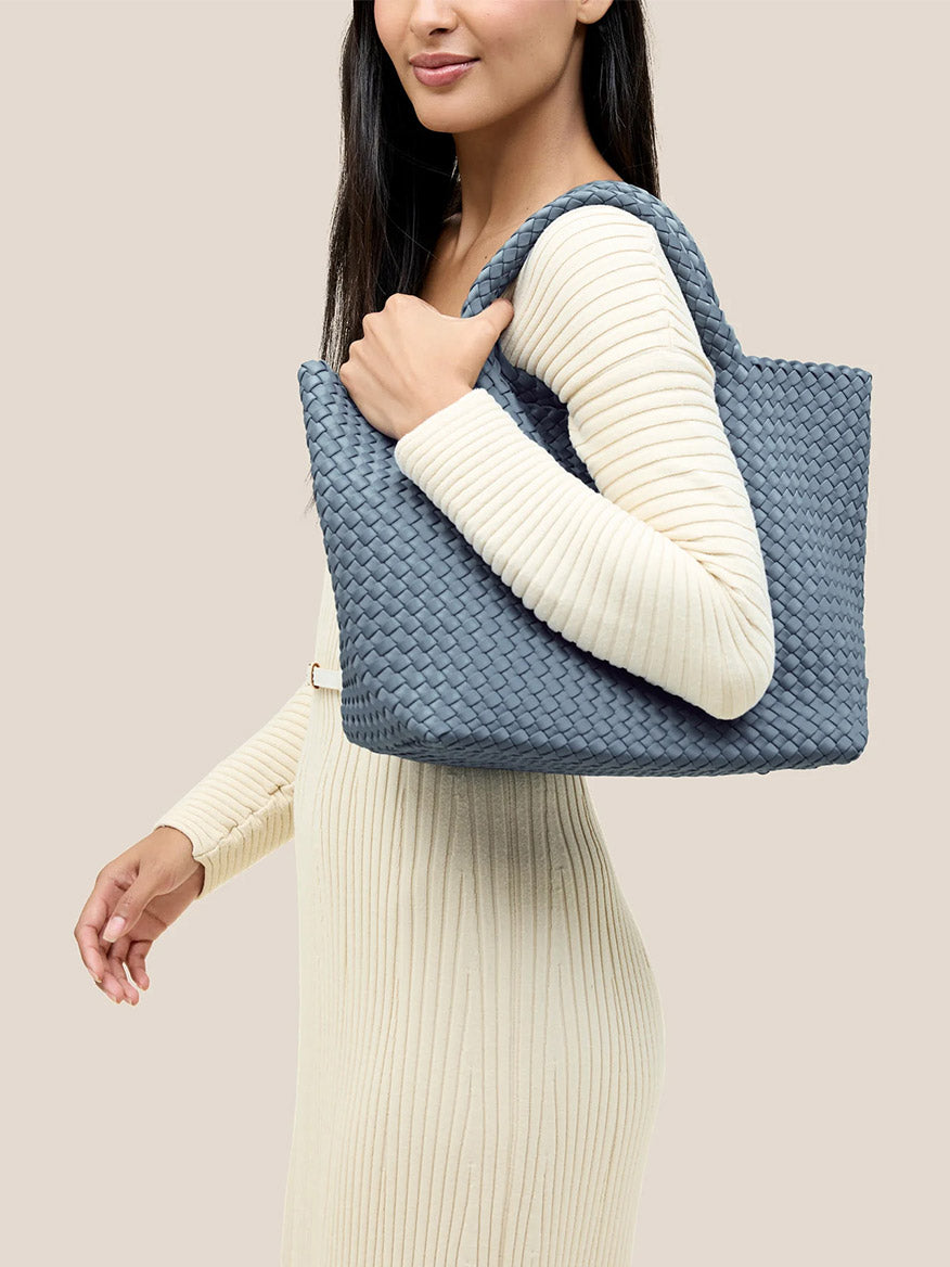 A person in a ribbed beige dress is holding a Naghedi St. Barths Medium Tote in Solid Nova, crafted from handwoven neoprene, over their shoulder.