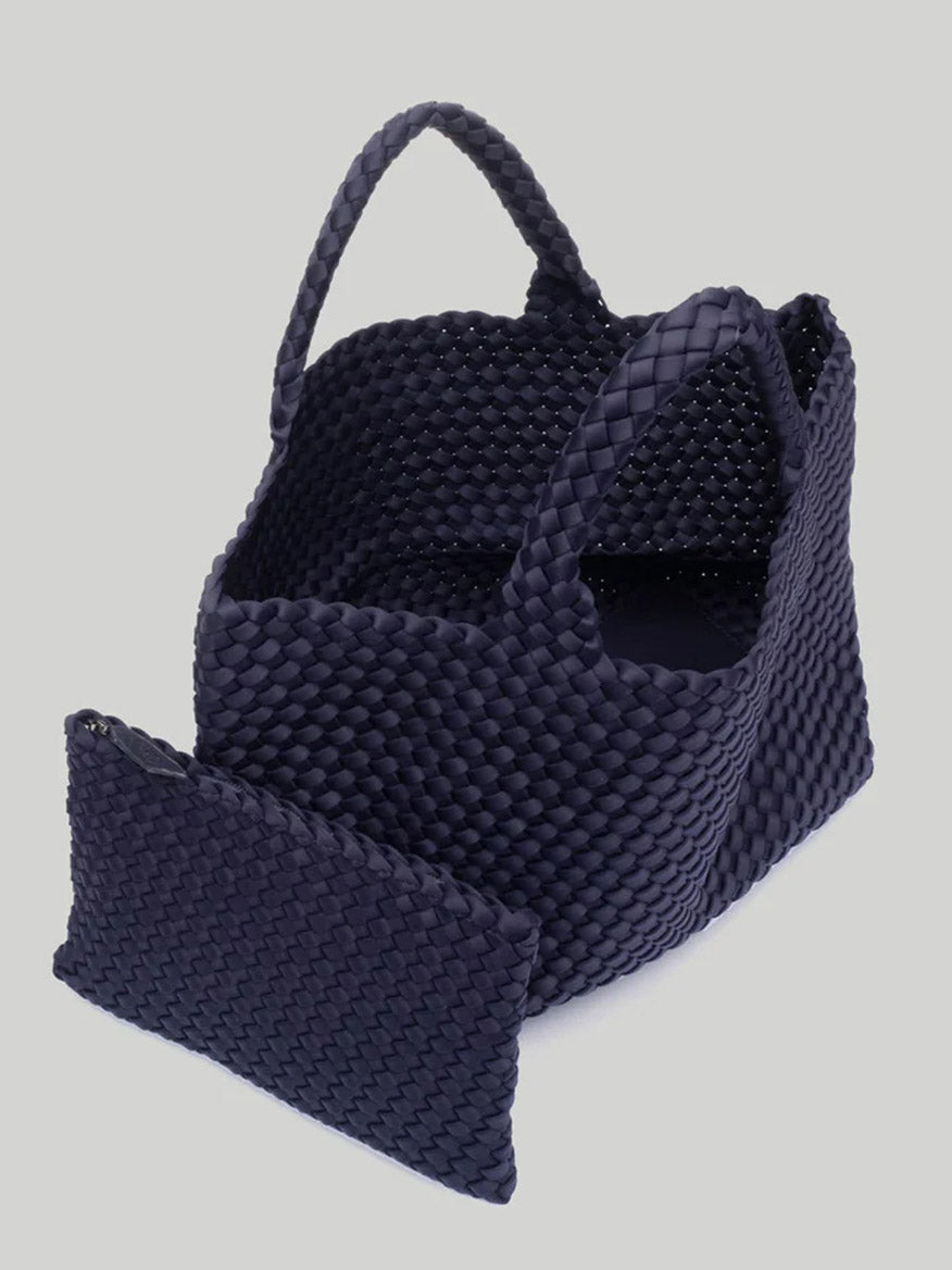 The Naghedi St. Barths Medium Tote in Solid Ink Blue, featuring a dark, handwoven neoprene design with two handles, is paired with a matching woven zippered pouch and displayed against a plain background.