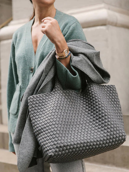 A person holds a fashionable Naghedi St. Barths Medium Tote in Solid Ink Blue and a gray coat, elegantly paired with a green cardigan. Their look is completed with a watch featuring a classic brown strap.