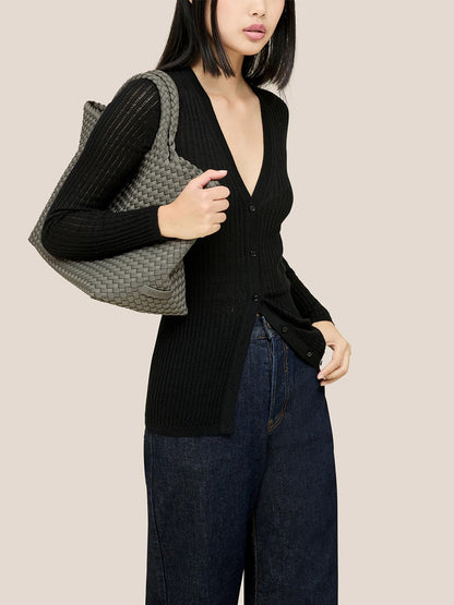 Dressed in a black cardigan and dark jeans, a person carries the Naghedi St. Barths Medium Tote in Solid Laurel, highlighting its ideal combination of style and functionality with its handwoven neoprene design.