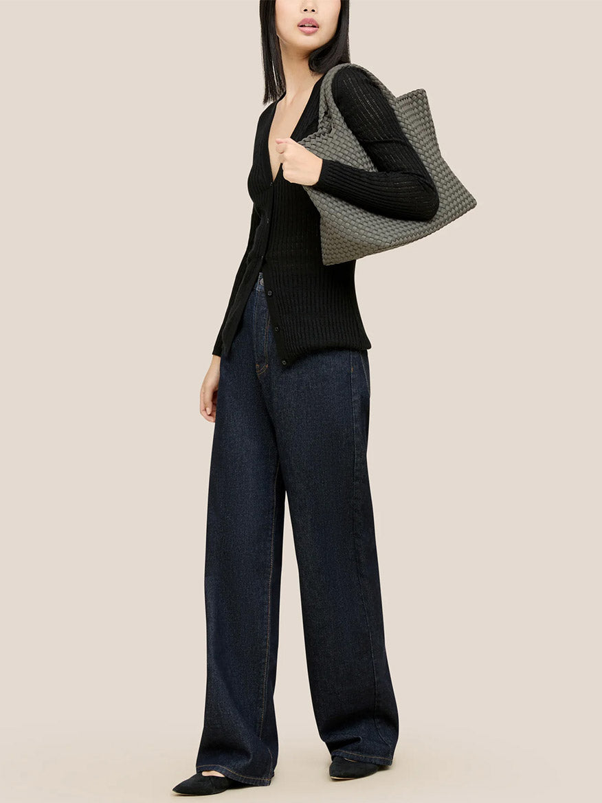 A person in a black cardigan and wide-leg jeans is carrying a versatile everyday bag, the Naghedi St. Barths Medium Tote in Solid Laurel, crafted from handwoven neoprene, over their shoulder.