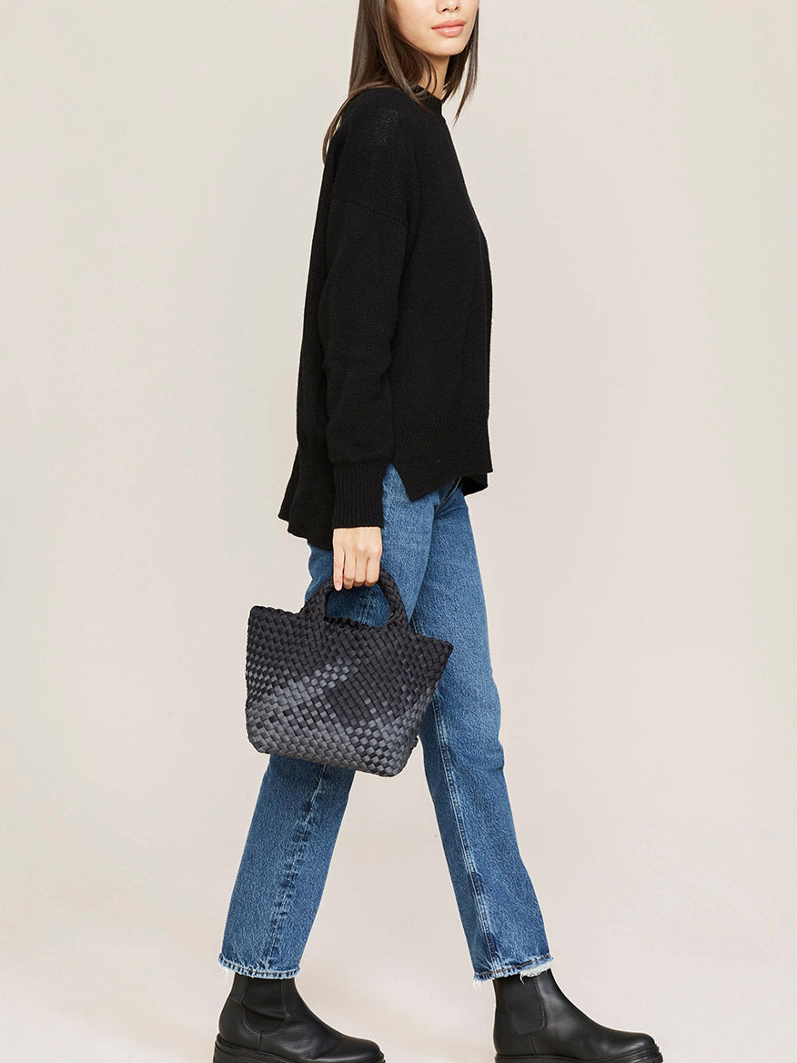 Side view of a woman walking, wearing a black sweater, blue jeans, and black boots, holding a Naghedi St. Barths Small Tote in Graphic Ombre Basalt.