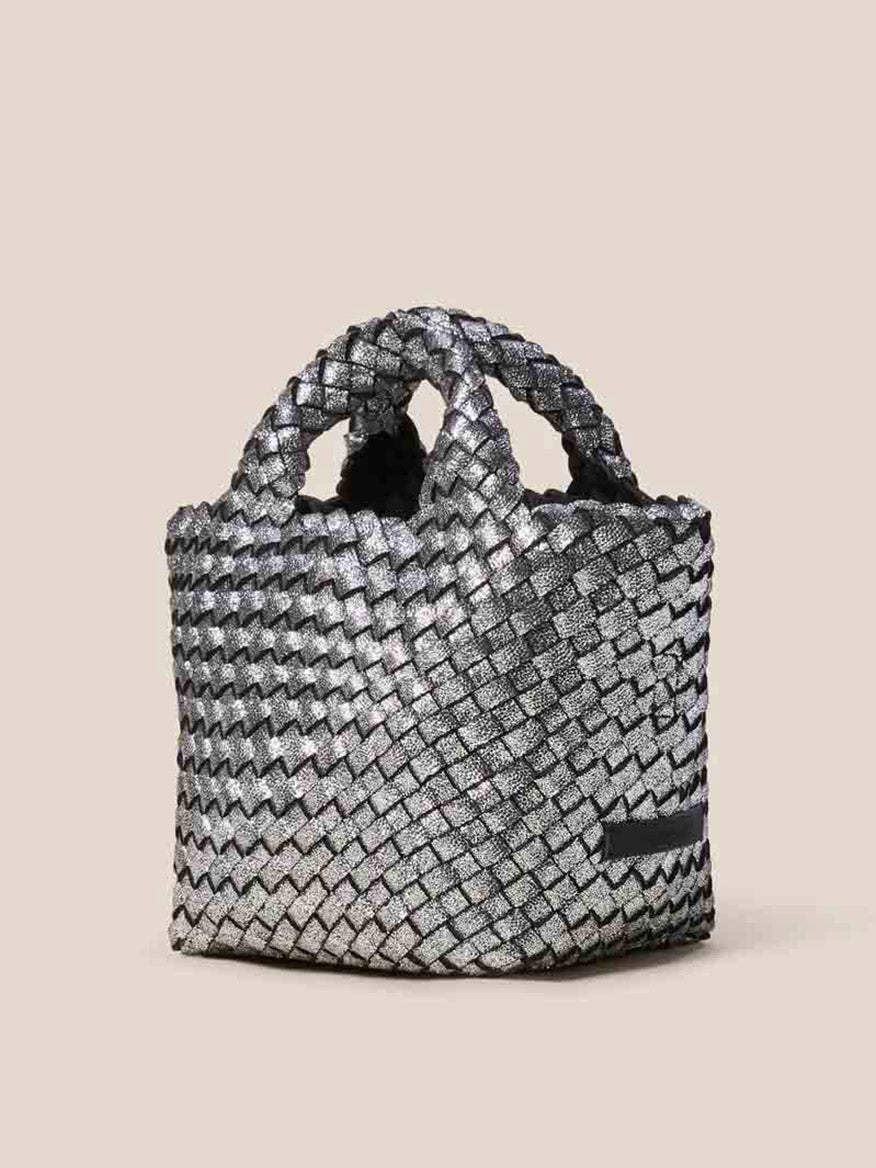 The Naghedi St. Barths Petit Tote in Metallic Titanium is a handbag crafted from handwoven neoprene with a textured pattern, featuring short handles. Set against a plain background, it offers an optional crossbody strap for versatile styling.