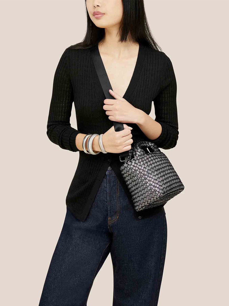 A person dressed in a black sweater and jeans carries a Naghedi St. Barths Petit Tote in Metallic Titanium, showcasing its unique handwoven neoprene design over their shoulder.