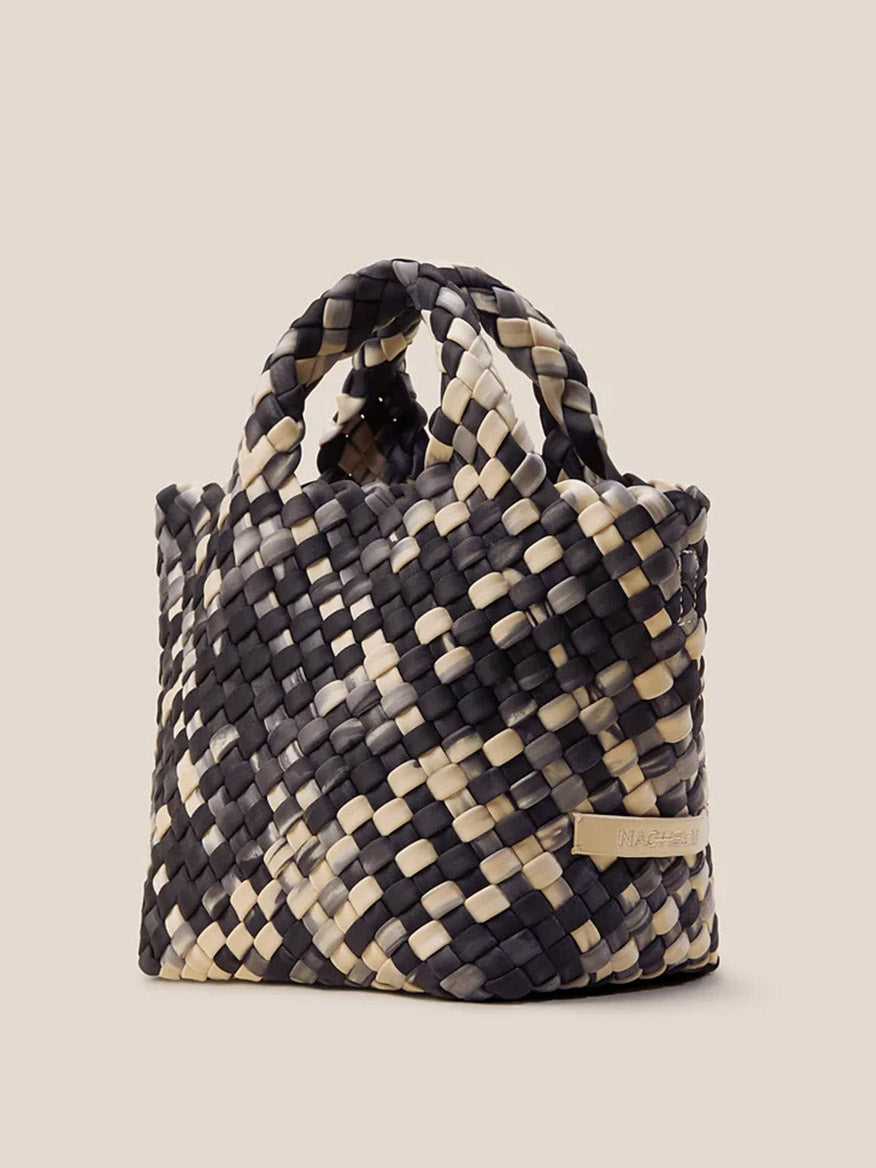 The Naghedi St. Barths Petit Tote in Watercolor Nimbus is beautifully highlighted against a neutral background with its handwoven neoprene design showcasing a black and beige checkered pattern. This versatile piece includes dual handles for easy carrying and a detachable crossbody strap for added convenience.