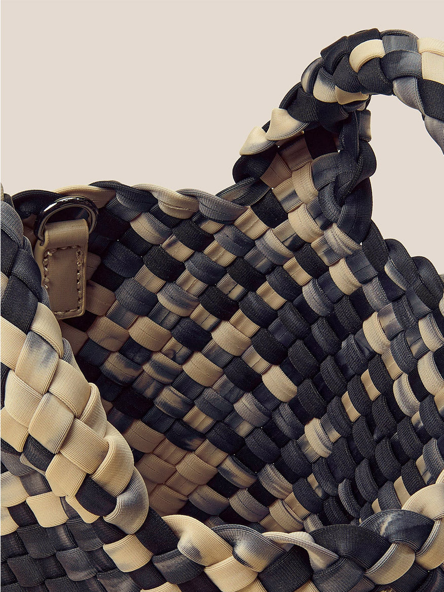 The Naghedi St. Barths Petit Tote in Watercolor Nimbus is a woven handbag that boasts a wide opening and a braided design, crafted from handwoven neoprene in black and beige tones. It features a detachable crossbody strap for enhanced versatility.