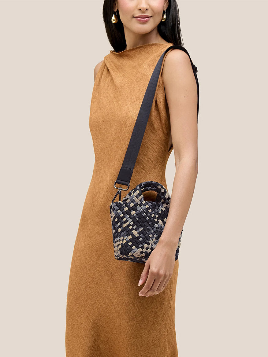 A woman in a brown dress elegantly holds the Naghedi St. Barths Petit Tote in Watercolor Nimbus, showcasing its handwoven neoprene design and detachable crossbody strap for versatile styling.