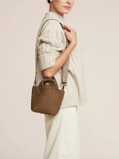 A person wearing a beige sweater and light-colored pants carries a small, brown, woven Naghedi St. Barths Petit Tote in Solid Mink with a crossbody strap over their shoulder.